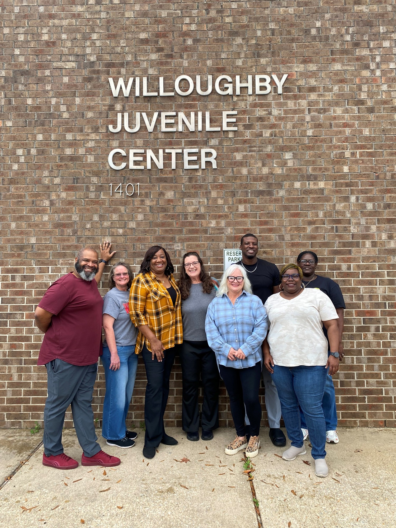 Group of eight staff members standing outside in front of a brick wall with the sign “Willoughby Juvenile Center”